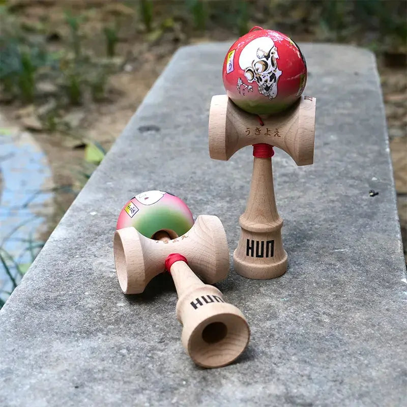 Two HUN kendamas with illustrated red-green balls, skull-style graphic artwork, and natural wood handles, displayed outdoors on a stone surface.