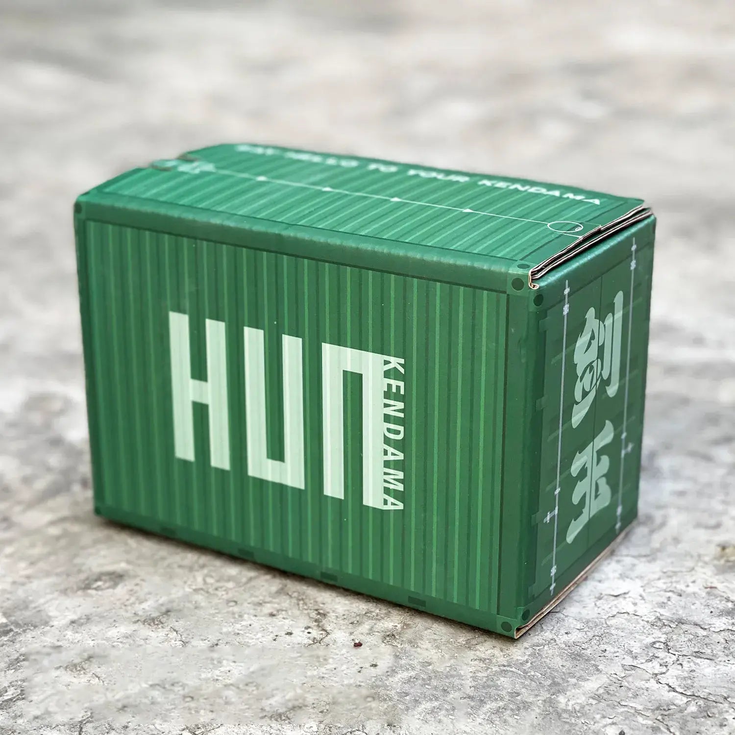 HUN Kendama green packaging box designed like a shipping container, displayed on a textured concrete surface.