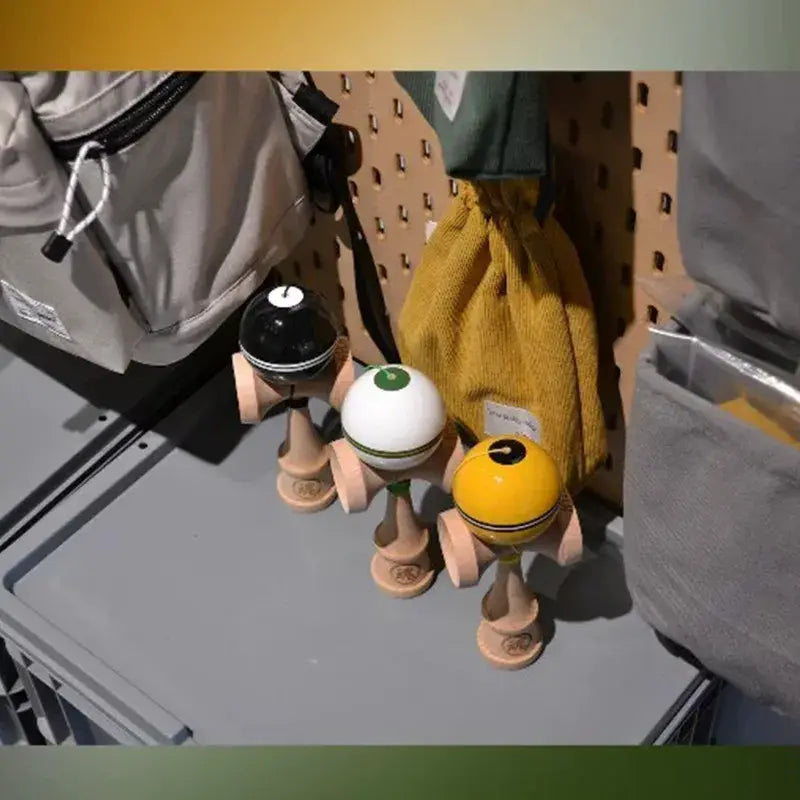 Three wooden kendamas with glossy black, white, and yellow balls, styled on a shelf next to bags and a mustard drawstring pouch.
