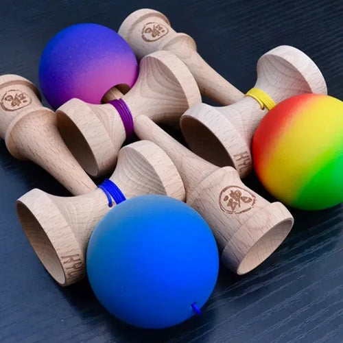 A group of wooden kendamas with colorful balls in blue, purple-pink, and rainbow gradient finishes, arranged on a dark surface.