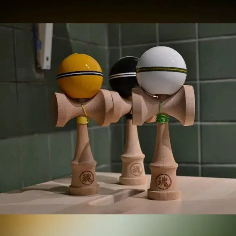 Three wooden kendamas with glossy yellow, black, and white balls, displayed together on a sink with a green tiled background.