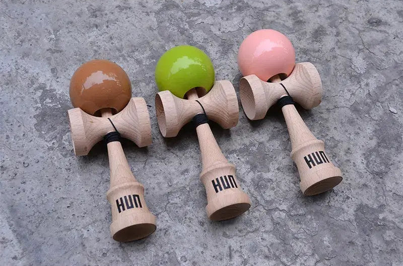 Three HUN kendamas with glossy brown, lime green, and pink balls and natural wood handles, displayed side by side on a textured concrete surface.