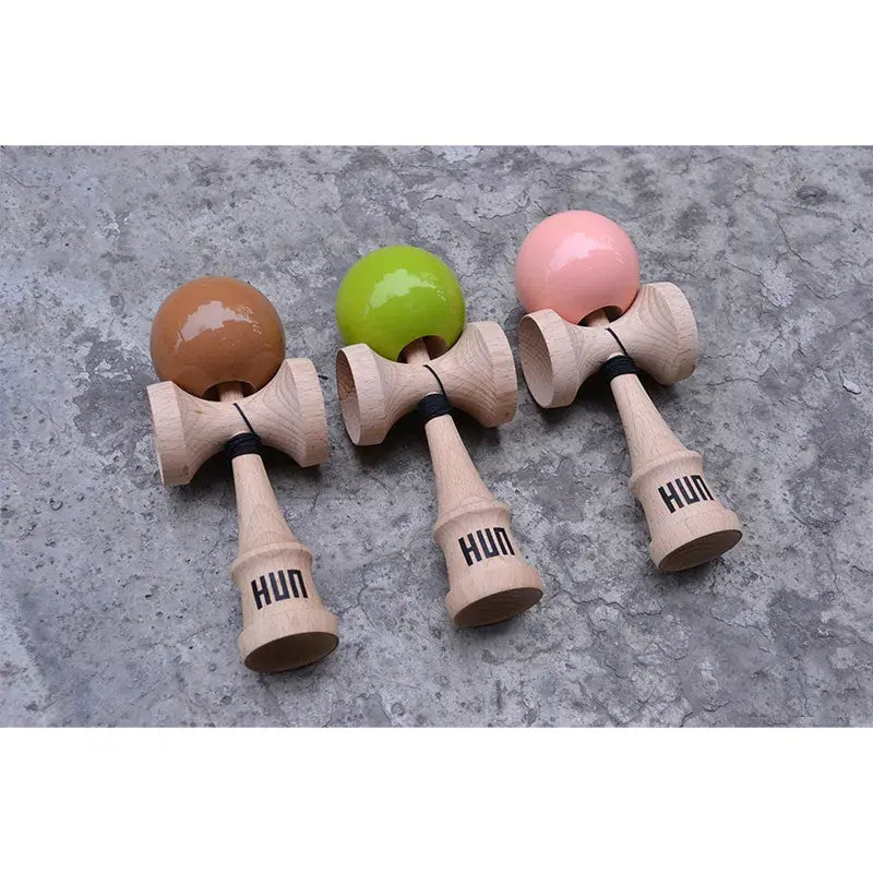 Three HUN kendamas with glossy brown, lime green, and pink balls and natural wood handles, displayed side by side on a textured concrete surface.