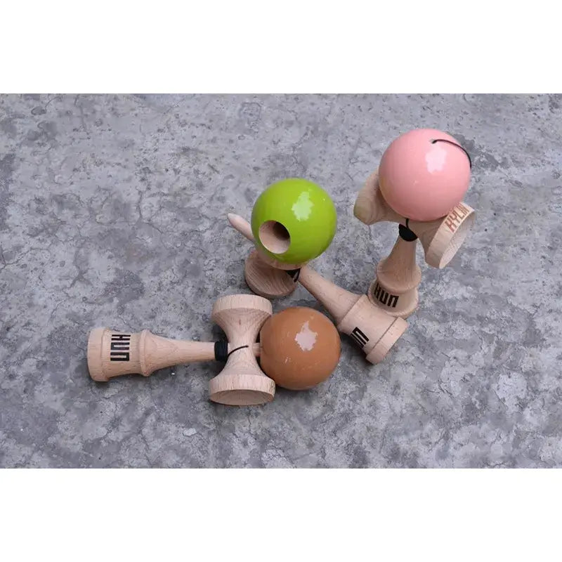 Three HUN kendamas with glossy brown, lime green, and pink balls and natural wood handles, arranged on a textured concrete surface in a top-down view.