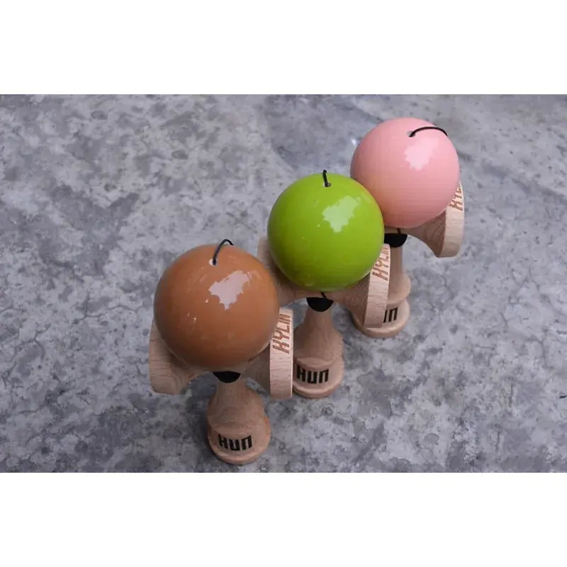 Three HUN kendamas with glossy brown, lime green, and pink balls and natural wood handles, displayed together on a textured concrete surface in a top-down view.