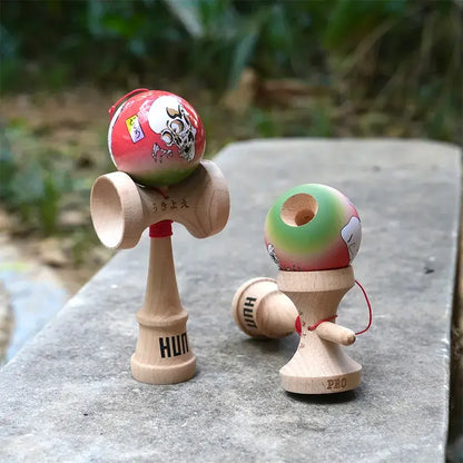 Two HUN kendamas with illustrated red-green balls, skull-style graphic artwork, and natural wood handles, displayed outdoors on a stone surface.