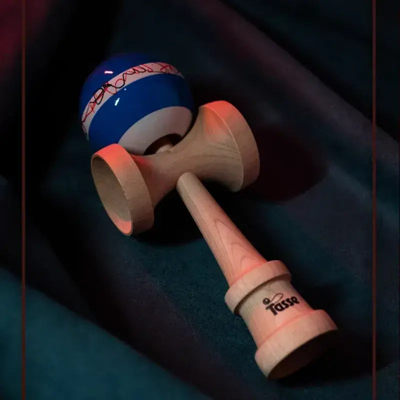 Tasse wooden kendama with a glossy blue ball, white band, red line graphic detail, and natural wood handle, styled diagonally on dark blue fabric.