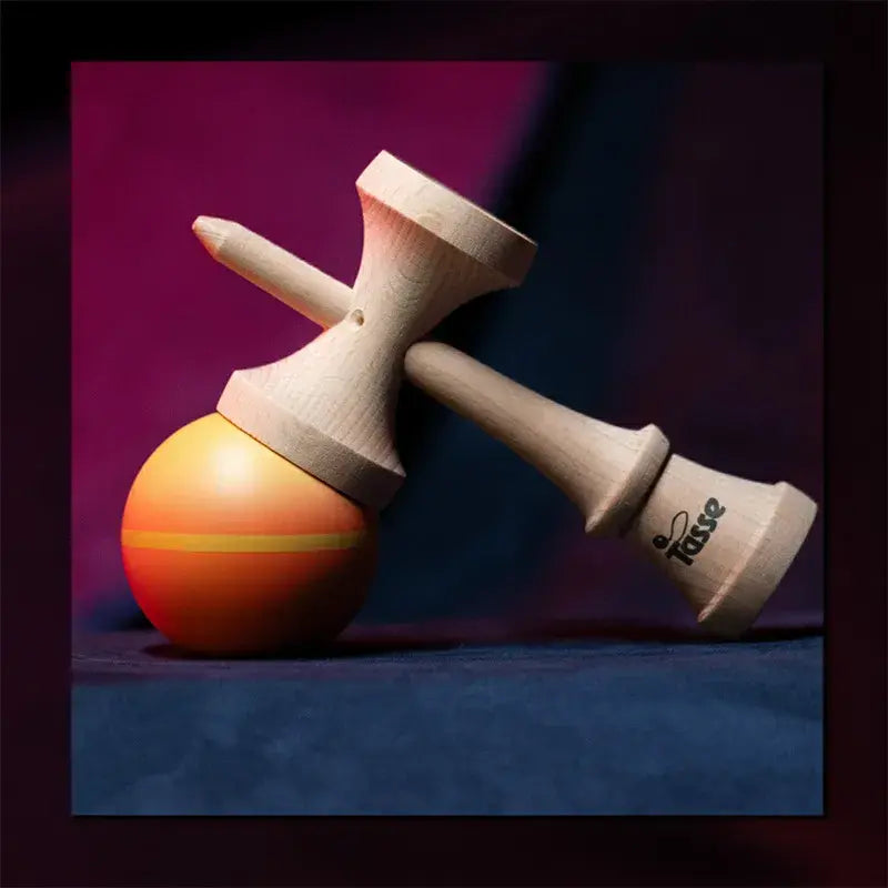 Tasse wooden kendama with a natural wood handle and orange-yellow gradient ball, styled diagonally on a dark red and blue background.