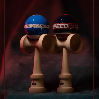 Two Tasse wooden kendamas with graphic blue and navy balls, including line art detail and “SPEED” text, displayed side by side on a dark red and blue background.