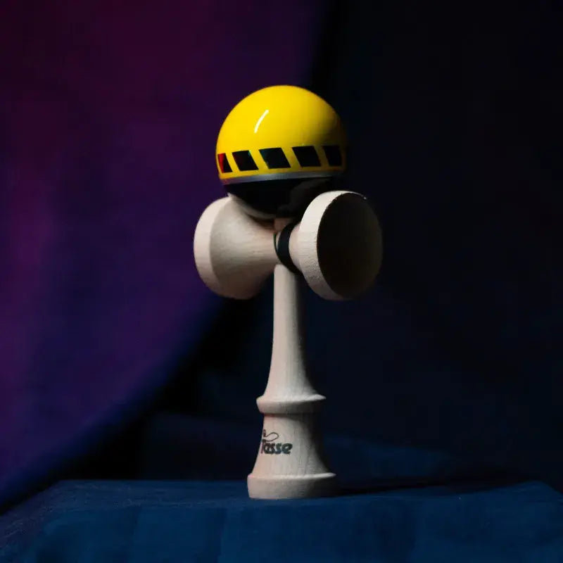 Tasse wooden kendama with a glossy yellow and black ball, striped graphic detail, and natural wood handle, displayed upright on a dark purple and blue background.