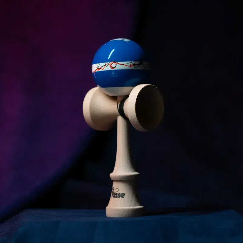 Tasse wooden kendama with a glossy blue ball, white band, red-black graphic detail, and natural wood handle, displayed upright on a dark purple and blue background.