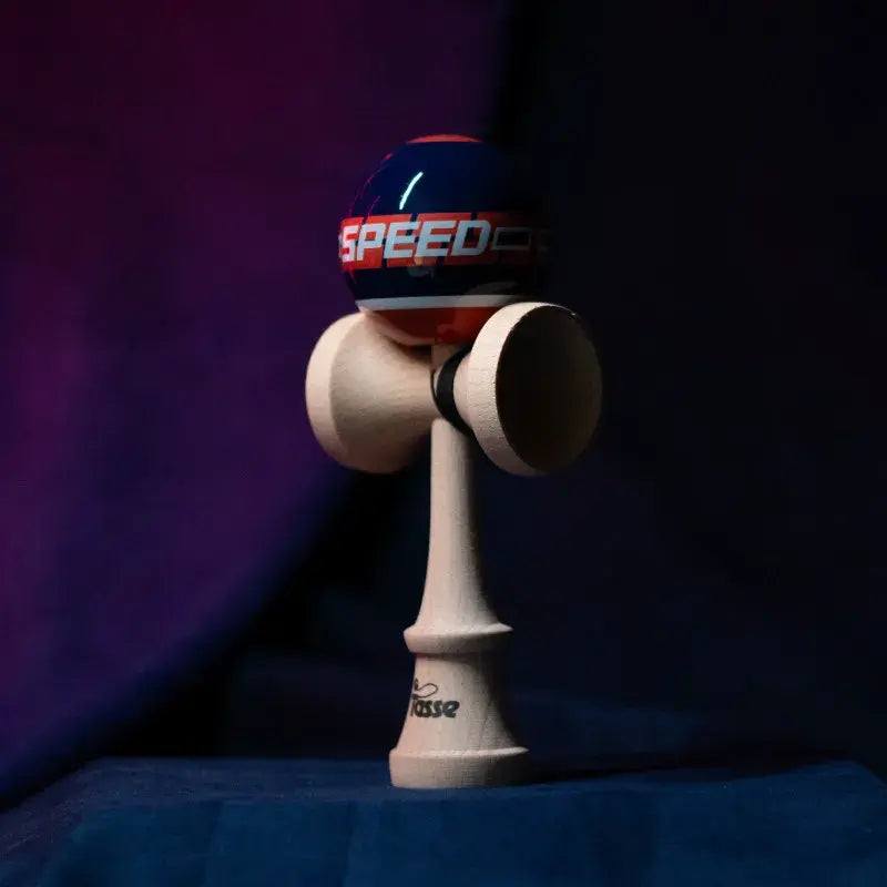 Tasse wooden kendama with a glossy navy and red ball, “SPEED” graphic detail, and natural wood handle, displayed upright on a dark purple and blue background.