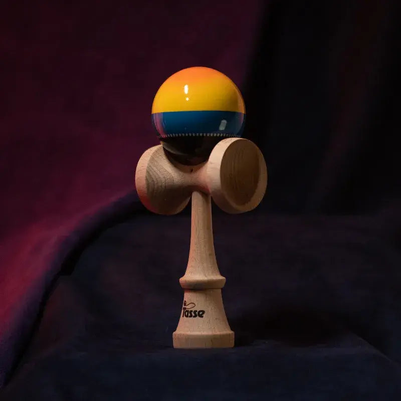 Tasse wooden kendama with a glossy yellow, blue, and black ball and natural wood handle, displayed upright on a dark red and blue background.