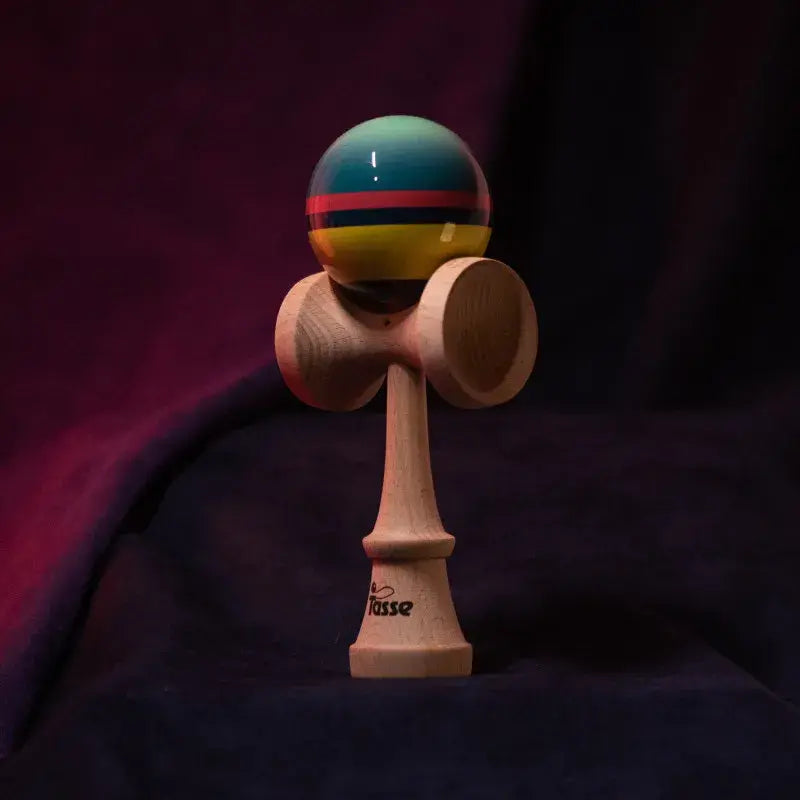 Tasse wooden kendama with a glossy teal, pink, yellow, and black striped ball and natural wood handle, displayed upright on a dark red and blue background.