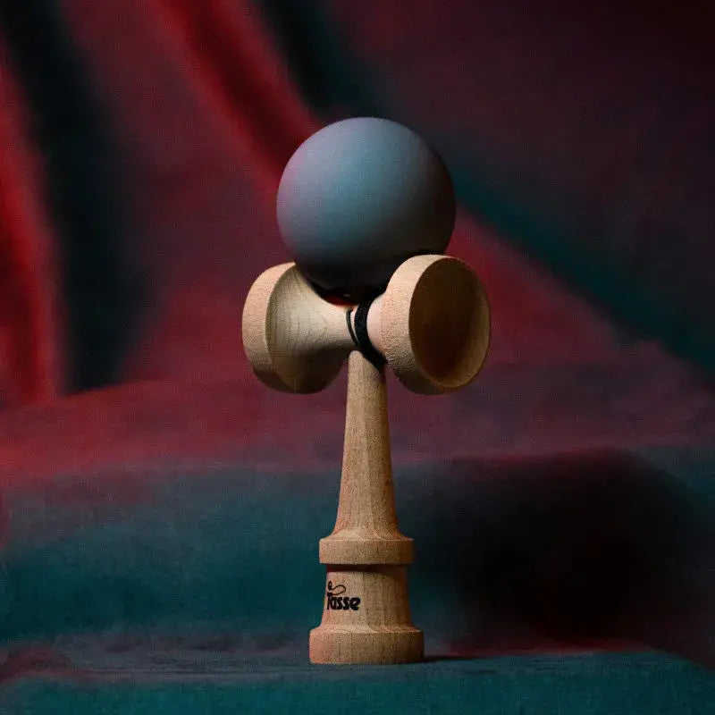 Tasse wooden kendama with a matte gray ball and natural wood handle, displayed upright on a red and blue gradient background.