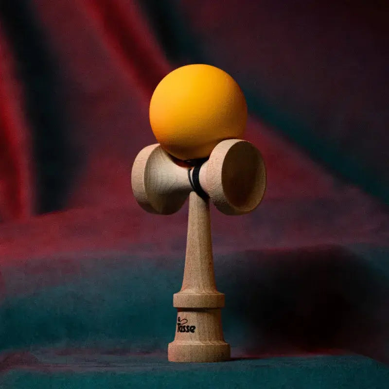 Tasse wooden kendama with a matte yellow ball and natural wood handle, displayed upright on a red and blue gradient background.