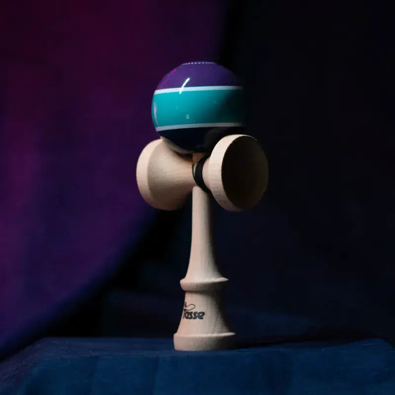 Tasse wooden kendama with a glossy purple and teal ball, white stripe detail, and natural wood handle, displayed upright on a dark purple and blue background.