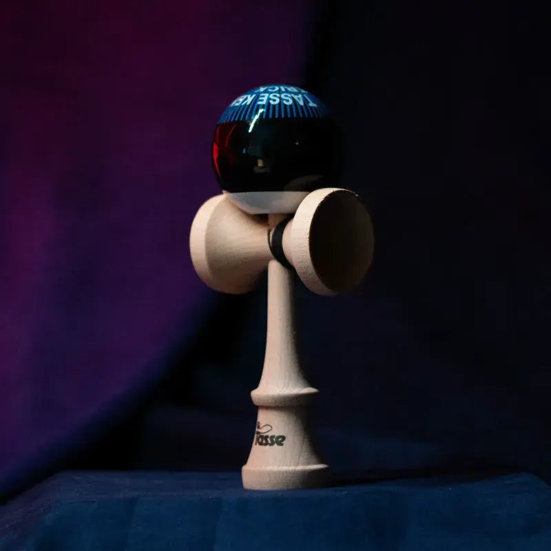Tasse wooden kendama with a glossy black ball, blue graphic text detail, and natural wood handle, displayed upright on a dark purple and blue background.
