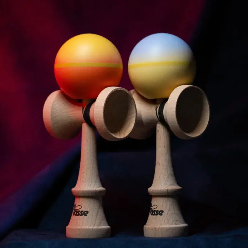 Tasse wooden kendama set with natural wood handles and orange-yellow and white-yellow gradient balls, displayed side by side on a dark red and blue background.