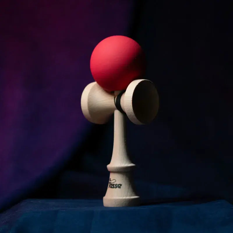 Tasse wooden kendama with a bright red ball and natural wood handle, displayed upright on a dark purple and blue background.