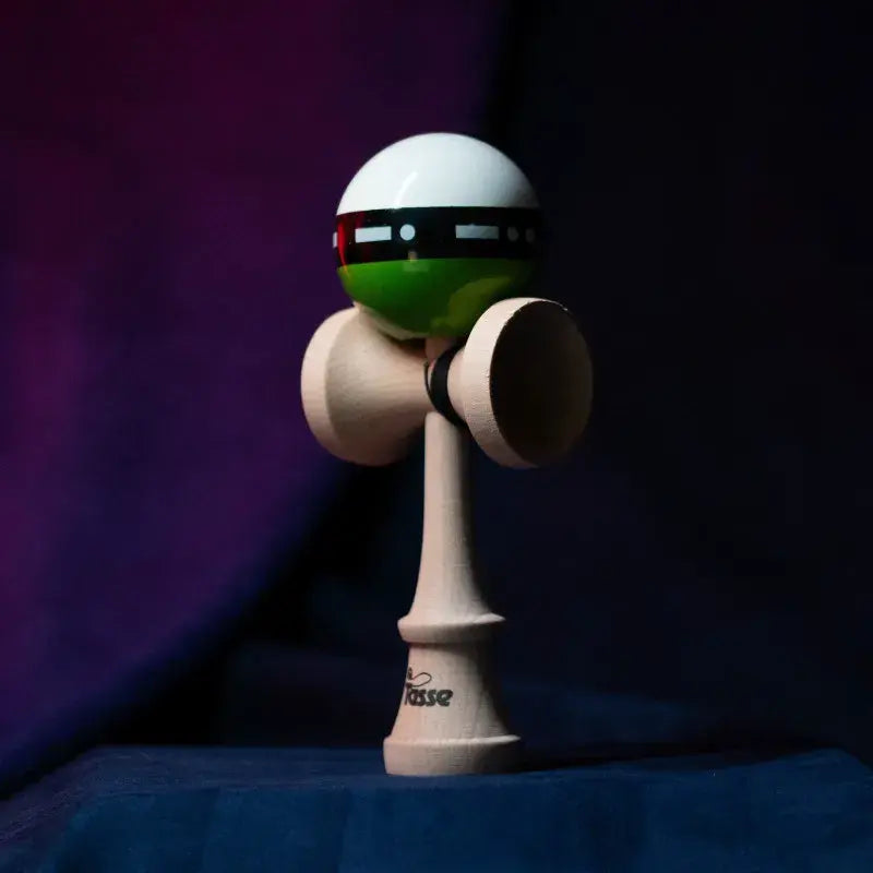 Tasse wooden kendama with a glossy white and green ball, black graphic band detail, and natural wood handle, displayed upright on a dark purple and blue background.
