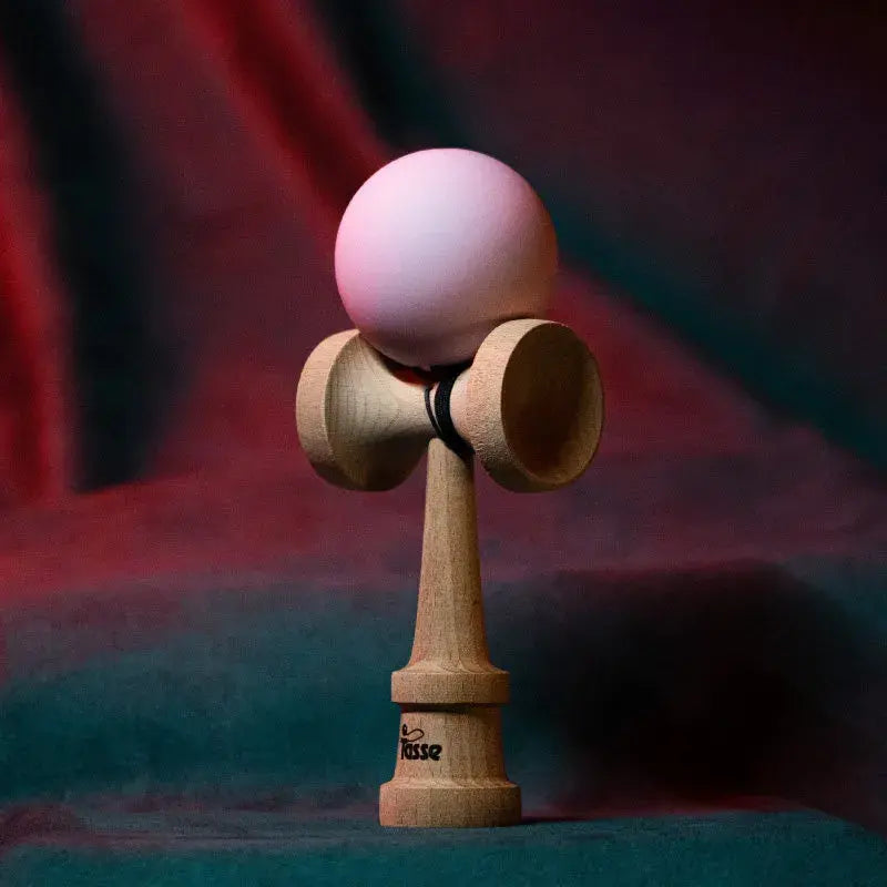 Tasse wooden kendama with a matte light pink ball and natural wood handle, displayed upright on a red and blue gradient background.