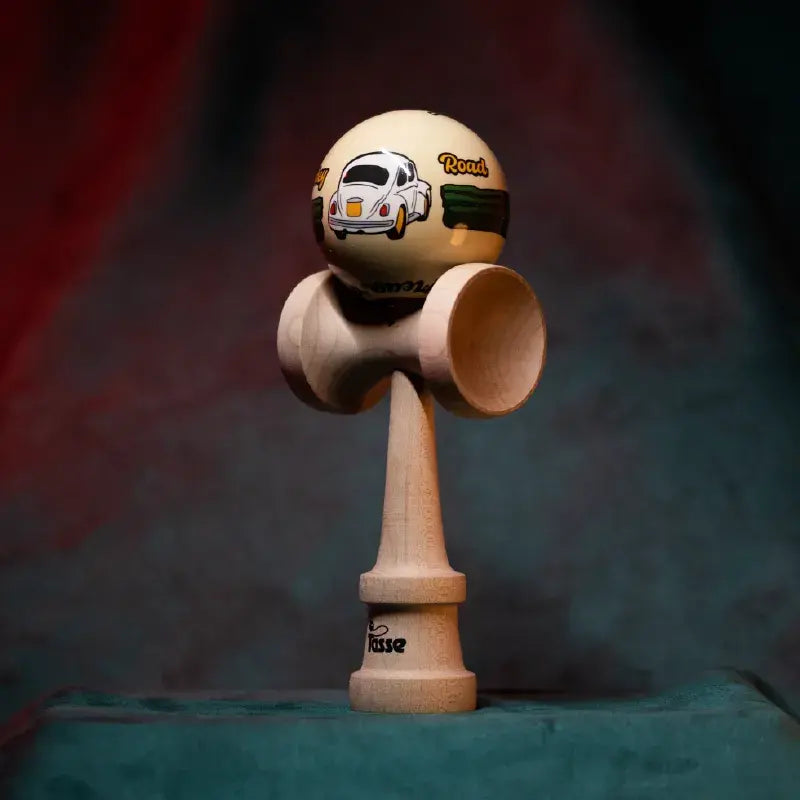 Tasse wooden kendama with a cream-colored ball, cartoon car illustration, “Road” graphic detail, and natural wood handle, displayed upright on a soft red and blue gradient background.