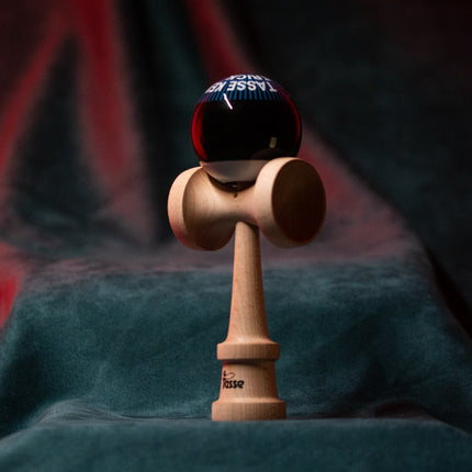 Tasse wooden kendama with a glossy black ball, blue graphic text detail, and natural wood handle, displayed upright on a red and blue background.