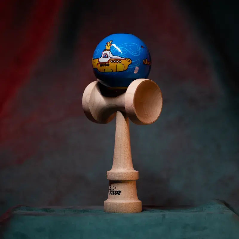 Tasse wooden kendama with a blue ball, yellow submarine graphic detail, and natural wood handle, displayed upright on a soft red and blue gradient background.