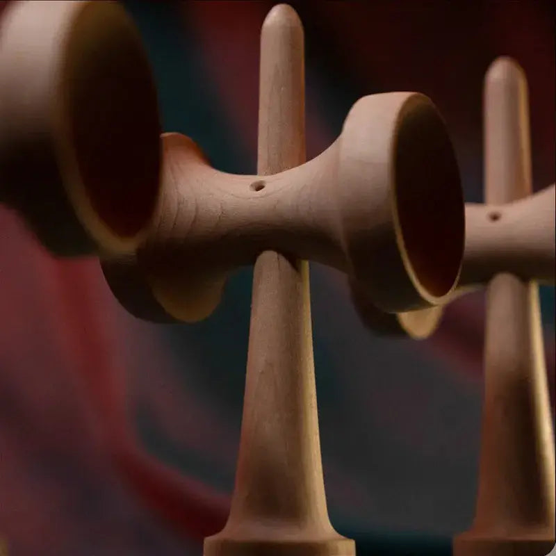 Close-up of a Tasse wooden kendama handle and cup details, with another kendama blurred in the background against a red and blue backdrop.